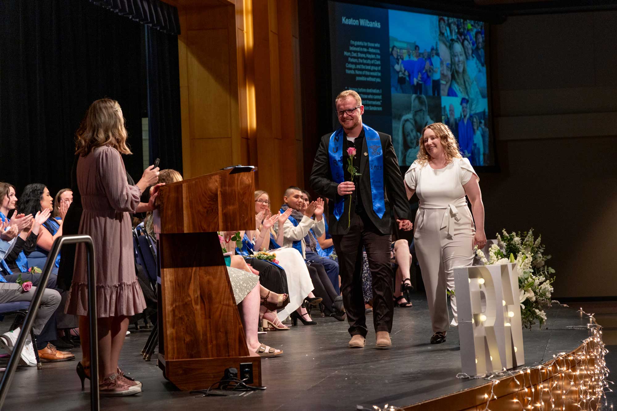 A nursing graduate walking down the stage with a companion, smiling and holding a single rose.